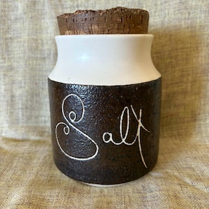 May include: A ceramic salt cellar with a cork stopper. The jar is white at the top and dark brown on the bottom, with the word "salt" written in white script. The jar is sitting on a beige surface.