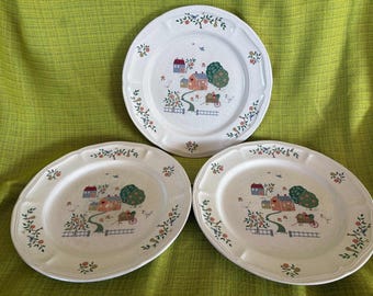 May include: Three white ceramic plates with a country scene design. Each plate features a house, tree, and cart illustration in the center, surrounded by a floral and fruit border. The plates have a scalloped edge.