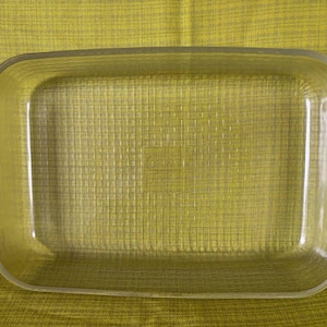 Puede incluir: Una fuente rectangular de vidrio transparente con esquinas redondeadas y asas. La fuente tiene un patrón texturizado en la parte inferior y está sobre un fondo amarillo-verdoso. Ideal para hornear guisos y otros platos.