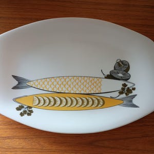 Op de afbeelding: Witte ovale serveerschaal met een geel en groen visontwerp. De vissen zijn gestileerd en hebben een herhalend schubbenpatroon. De schaal heeft een witte achtergrond met een subtiele glans.