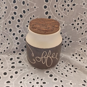 May include: A brown ceramic coffee jar with a white top and a cork lid. The jar has the word "coffee" written on it in white.
