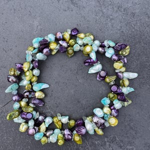 May include: A multi-strand necklace composed of irregular, baroque-style pearls in a vibrant colour palette. The pearls feature iridescent shades of deep purple, lime green, turquoise, and pale blue, arranged in a clustered, organic circular formation.