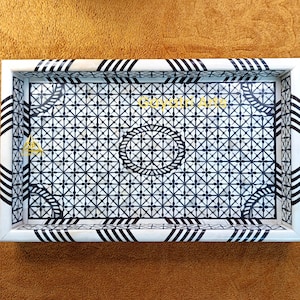 Könnte beinhalten: Rechteckiges dekoratives Tablett mit weißem Rahmen und einem schwarz-weißen geometrischen Muster. Das Innere des Tabletts weist ein sich wiederholendes Kreuz- und Kreisdesign auf. Der Rahmen hat schwarz-weiße Streifenakzente. Das Tablett liegt auf einer braunen Oberfläche.