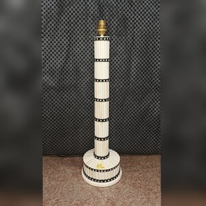 Op de afbeelding: Een hoge, cilindrische lampvoet met een gecanneleerde witte keramische kolom, geaccentueerd door horizontale zwarte banden met witte stippen. De voet heeft een ronde, gecanneleerde sokkel en een messingkleurige fitting aan de bovenzijde.