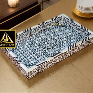 May include: Decorative rectangular tray with a geometric black and white pattern. The tray has a white border with black accents and a central circular design. The tray is on a gold-colored surface.