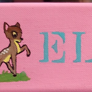 K&ouml;nnte beinhalten: Ein rosa Canvas-Schild mit dem Namen "ELENA", der in hellblau geschrieben ist. Das Schild ist mit zwei Comic-Hirschen auf beiden Seiten des Namens verziert.