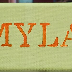 K&ouml;nnte beinhalten: Ein gr&uuml;nes Leinwand-Schild mit dem Namen "MYLA", der in orangefarbenen Buchstaben geschrieben ist. Zwei braune Eichh&ouml;rnchen mit wei&szlig;em Bauch sind auf beiden Seiten des Namens gemalt.