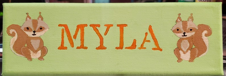 K&ouml;nnte beinhalten: Ein gr&uuml;nes Leinwand-Schild mit dem Namen "MYLA", der in orangefarbenen Buchstaben geschrieben ist. Zwei braune Eichh&ouml;rnchen mit wei&szlig;em Bauch sind auf beiden Seiten des Namens gemalt.