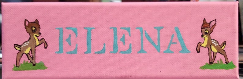 K&ouml;nnte beinhalten: Ein rosa Canvas-Schild mit dem Namen "ELENA", der in hellblau geschrieben ist. Das Schild ist mit zwei Comic-Hirschen auf beiden Seiten des Namens verziert.