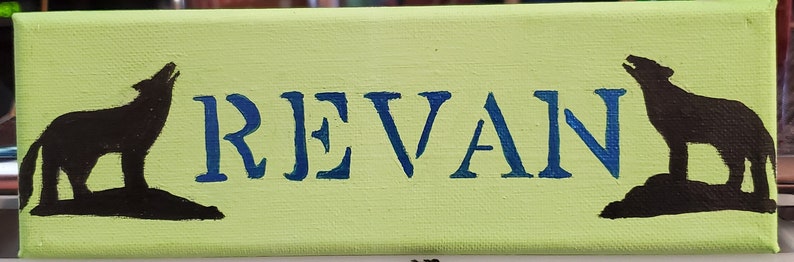 K&ouml;nnte beinhalten: Ein hellgr&uuml;nes Leinwand-Schild mit dem Namen "REVAN" in blauer Farbe. Zwei schwarze Wolf-Silhouetten befinden sich auf beiden Seiten des Namens.