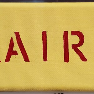 K&ouml;nnte beinhalten: Ein gelbes Leinwand-Schild mit dem Namen "KAIRI" in roten Buchstaben. Zwei schwarz-wei&szlig;e Mickey-Maus-Figuren sind auf beiden Seiten des Namens gemalt.