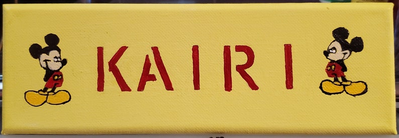K&ouml;nnte beinhalten: Ein gelbes Leinwand-Schild mit dem Namen "KAIRI" in roten Buchstaben. Zwei schwarz-wei&szlig;e Mickey-Maus-Figuren sind auf beiden Seiten des Namens gemalt.