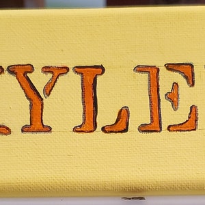 K&ouml;nnte beinhalten: Eine gelbe Leinwand mit dem Namen "KYLEE" in orangefarbenen Buchstaben. Zwei braune Faultiere sind auf beiden Seiten des Namens gemalt.