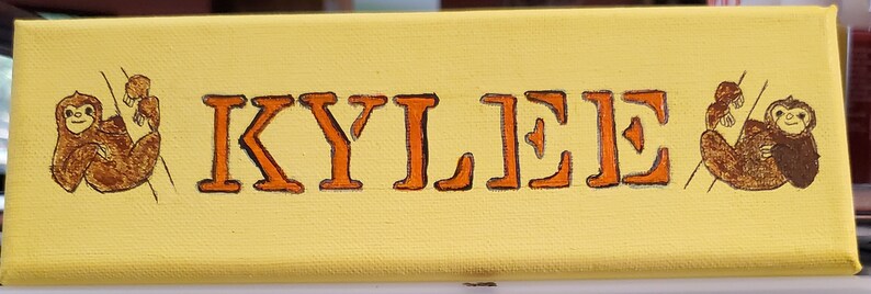 K&ouml;nnte beinhalten: Eine gelbe Leinwand mit dem Namen "KYLEE" in orangefarbenen Buchstaben. Zwei braune Faultiere sind auf beiden Seiten des Namens gemalt.
