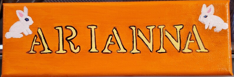 K&ouml;nnte beinhalten: Ein orangefarbenes Leinwand-Schild mit dem Namen "ARIANNA", der in gelben Buchstaben geschrieben ist. Zwei wei&szlig;e Kaninchen sind auf beiden Seiten des Namens gemalt.