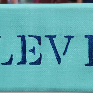 K&ouml;nnte beinhalten: Ein t&uuml;rkisfarbenes Leinwand-Schild mit zwei grauen Elefanten mit rosa Ohren und wei&szlig;en Akzenten. Der Name "LEVI" ist in blauen Buchstaben gemalt.