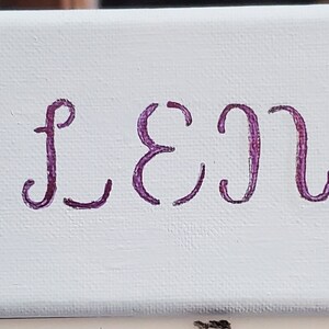 K&ouml;nnte beinhalten: Ein wei&szlig;es Leinwand-Schild mit dem Namen "ELENA" in lila Kursivschrift. Zwei schwarz-wei&szlig;e Pferdezeichnungen flankieren den Namen.