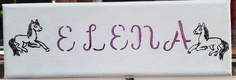 K&ouml;nnte beinhalten: Ein wei&szlig;es Leinwand-Schild mit dem Namen "ELENA" in lila Kursivschrift. Zwei schwarz-wei&szlig;e Pferdezeichnungen flankieren den Namen.