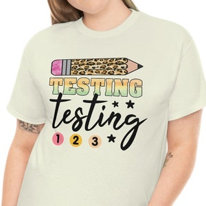 以下が含まれることがあります： レオパード柄の鉛筆のグラフィックがプリントされた薄いベージュのTシャツ。Tシャツには「TESTING testing 1 2 3」の文字がプリントされています。
