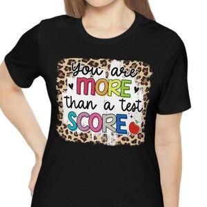 Könnte beinhalten: Schwarzes T-Shirt mit einem Leopardenmuster-Druck, auf dem "You are more than a test score" mit einem roten Apfel und Herzen steht.