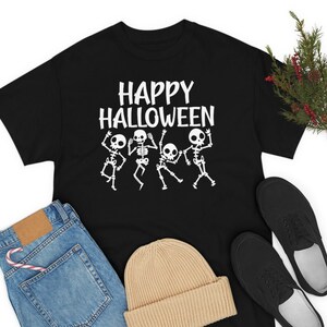 May include: Black t-shirt with the text "Happy Halloween" and a graphic of four dancing skeletons. The shirt is laid flat on a white surface with a pair of black shoes, a pair of blue jeans, and a beige beanie hat.