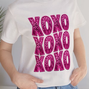 Puede incluir: Camiseta blanca con un diseño de purpurina rosa que dice "XOXO" tres veces.