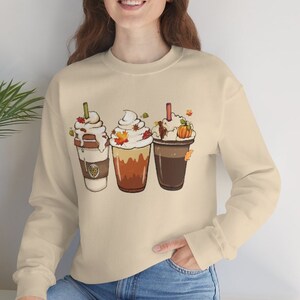Puede incluir: Una sudadera beige con un gráfico de tres bebidas de café de otoño con crema batida y hojas de otoño. Las bebidas están en una fila, de izquierda a derecha, y están coronadas con crema batida y hojas de otoño.