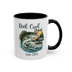 May include: White ceramic mug with a black interior and handle. The mug features a graphic of a fish jumping out of the water with the words "Reel Cool Dad" and "Since 2015". The design includes a fishing scene with trees and a boat.