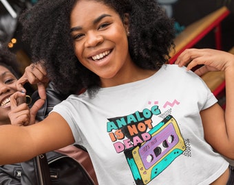 May include: White t-shirt with a colorful graphic of a cassette tape and the text "ANALOG IS NOT DEAD 90S HITS". The design features teal, purple, pink, and yellow accents. The shirt is worn by a person.