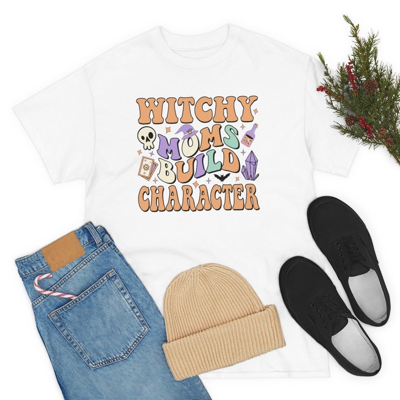 May include: White t-shirt with the text "Witchy Moms Build Character" in orange and purple letters. The text is surrounded by various witchy icons, including a skull, a witch's hat, a potion bottle, a tarot card, a bat, and a crystal.