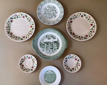 May include: Set of seven vintage ceramic plates with floral and house designs. The plates are white with green and red floral patterns and a green house design.