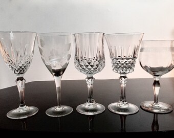 May include: Five clear glass wine glasses with different etched designs. The glasses are arranged in a row on a black surface.