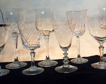 May include: A collection of eight clear glass wine glasses with various designs and shapes. The glasses are arranged on a black surface.