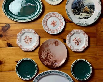 May include: A collection of nine decorative plates in various sizes and designs. The plates feature floral patterns, a winter scene, and green and white borders.