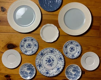 May include: A collection of 10 decorative plates in various sizes and designs. The plates feature blue and white floral patterns, some with a raised texture. The plates are arranged in a circular pattern on a wooden surface.
