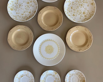 May include: A collection of cream-colored and tan ceramic plates and saucers. Some plates feature a floral design in gold, while others have a geometric pattern or a solid color. The arrangement includes various sizes, suitable for a dining set.