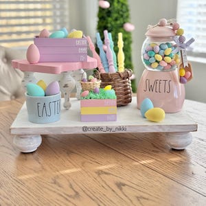 May include: A tiered wooden stand with pastel Easter eggs, a basket of pastel eggs, and a pink ceramic jar with the word "SWEETS" on it. The jar is filled with pastel candy eggs. The stand is decorated with the words "Easter Eggs 25¢" and "Easter Egg Hunt".