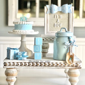 May include: A white tiered stand with blue and white decorations. The top tier has a blue and white cake with a blue 'BOY' topper. The middle tier has a blue and white 'BOY' block set. The bottom tier has a blue and white ceramic jar with a blue and white ribbon and a wooden spoon with a blue and white tag that says 'It's a Boy'.