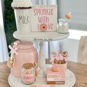 May include: A tiered tray decorated with a donut-themed design. The top tier features a white mug with the word "MILK" on it, a donut with sprinkles, and a sign that says "Sprinkle with Love". The bottom tier features a pink jar filled with sprinkles, a wooden spoon, and a sign that says "Life is better with sprinkles".