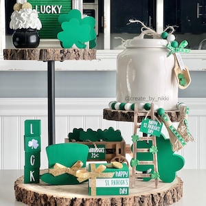 May include: A tiered tray with St. Patrick's Day decorations. Includes a green felt sign that says "LUCKY", a green top hat, wooden blocks spelling "LUCK", and a white jar. The tray is decorated with shamrocks and gold accents.