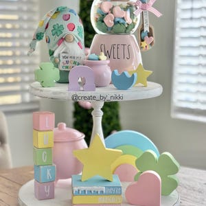 May include: A two-tiered white wooden tray with pastel-colored decorative items. Includes a gnome, a jar of sweets, blocks spelling "LUCKY", and various shapes like stars, hearts, and clovers. The text "They're magically delicious" is visible on a stack of books.