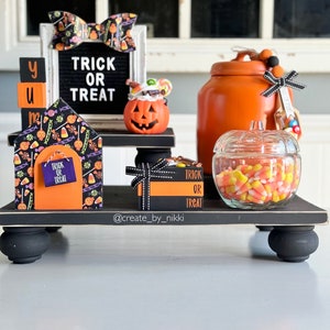 May include: A Halloween-themed display featuring a tiered black tray with various decorations. Includes a sign that says "Trick or Treat", a pumpkin-shaped candy holder, a jar of candy corn, and decorative blocks. The color scheme is orange, black, and white.