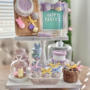 May include: A two-tiered white tiered tray decorated for Easter. The top tier features a wooden cutting board with a purple cake, a pink bunny, a yellow lollipop, and a purple macaron. The middle tier has a sign that says "Happy Easter" and a rolling pin that says "Peeps". The bottom tier has a glass cloche with a bunny cookie, a bowl of cake pops with sprinkles and yellow and blue bows, and a wicker basket.