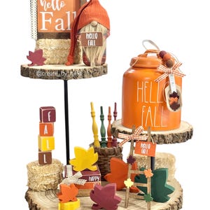 May include: A fall-themed decorative display featuring an orange jar with "HELLO FALL" text, a gnome with a "HELLO FALL" sign, wooden leaf cutouts, and a small ladder with a "HELLO FALL" sign. The display includes wooden blocks spelling "FALL".
