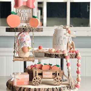 May include: A three-tiered wooden stand decorated with peach-themed decorations. The top tier features a gnome wearing a peach-patterned hat and swinging on a rope swing. The middle tier has a white ceramic sugar jar with the word "SUGAR" written on it. The bottom tier has a wooden wagon with the words "FRESH PEACHES" on it, a small wooden box with the words "SWEET AS A PEACH" on it, and a peach-colored beaded garland.