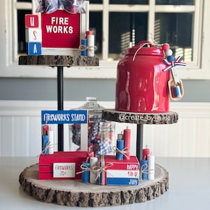 May include: A tiered wooden stand decorated for the Fourth of July. The top tier features a red letter board with the words "FIRE WORKS" and a small wooden block spelling "USA". The middle tier has a red ceramic jar with a white ribbon and a small wooden sign that says "FIREWORKS STAND". The bottom tier has a red wooden box with the words "FIREWORKS 25c" and a stack of small wooden blocks that say "HAPPY 4th OF JULY".