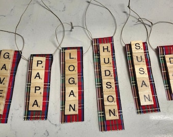 May include: Five Christmas decorations made from wooden letter tiles spelling out the names GAVEN, PAPA, LOGAN, HUDSON, and DAD. Each decoration is attached to a red and black tartan ribbon.