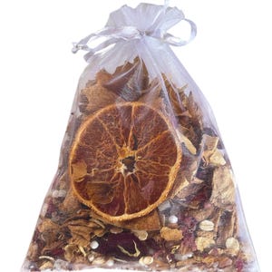 May include: A clear, sheer bag filled with dried botanicals, including a large, dried orange slice. The bag is tied with a white ribbon. The contents are a mix of brown, red, and white elements, suggesting a potpourri or sachet.
