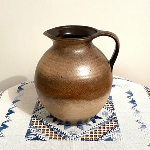 Pode incluir: Um jarro de cerâmica castanho com interior e alça castanhos escuros. O jarro tem um corpo arredondado e um bico. Está sobre uma toalha de mesa branca com padrões geométricos azuis.