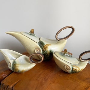 Może przedstawiać: Zestaw do herbaty z ceramiki w stylu vintage, składający się z czajniczka, dzbanka do śmietanki i cukiernicy. Zestaw ma kremowe szkliwo z brązowymi akcentami na uchwytach i pokrywkach. Ozdobne zielone i brązowe wzory kwiatowe zdobią boki.